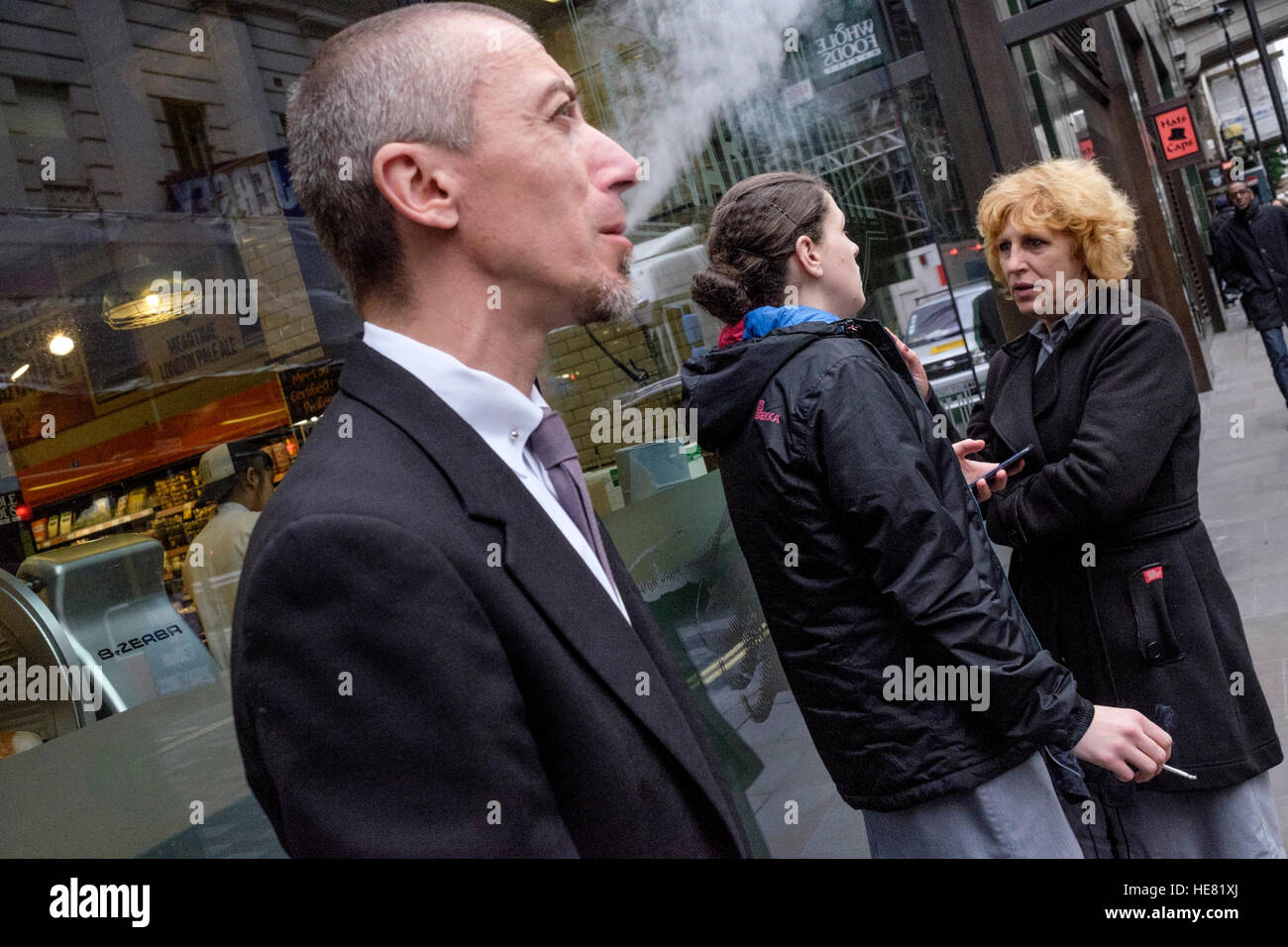 A man breathes out cigarette smoke whilst two women chat in the background. Stock Photo