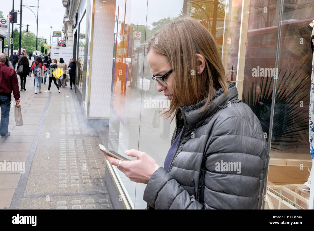 A young woman captured having a cigarette whilst using her mobile phone on one of London's busy streets Stock Photo