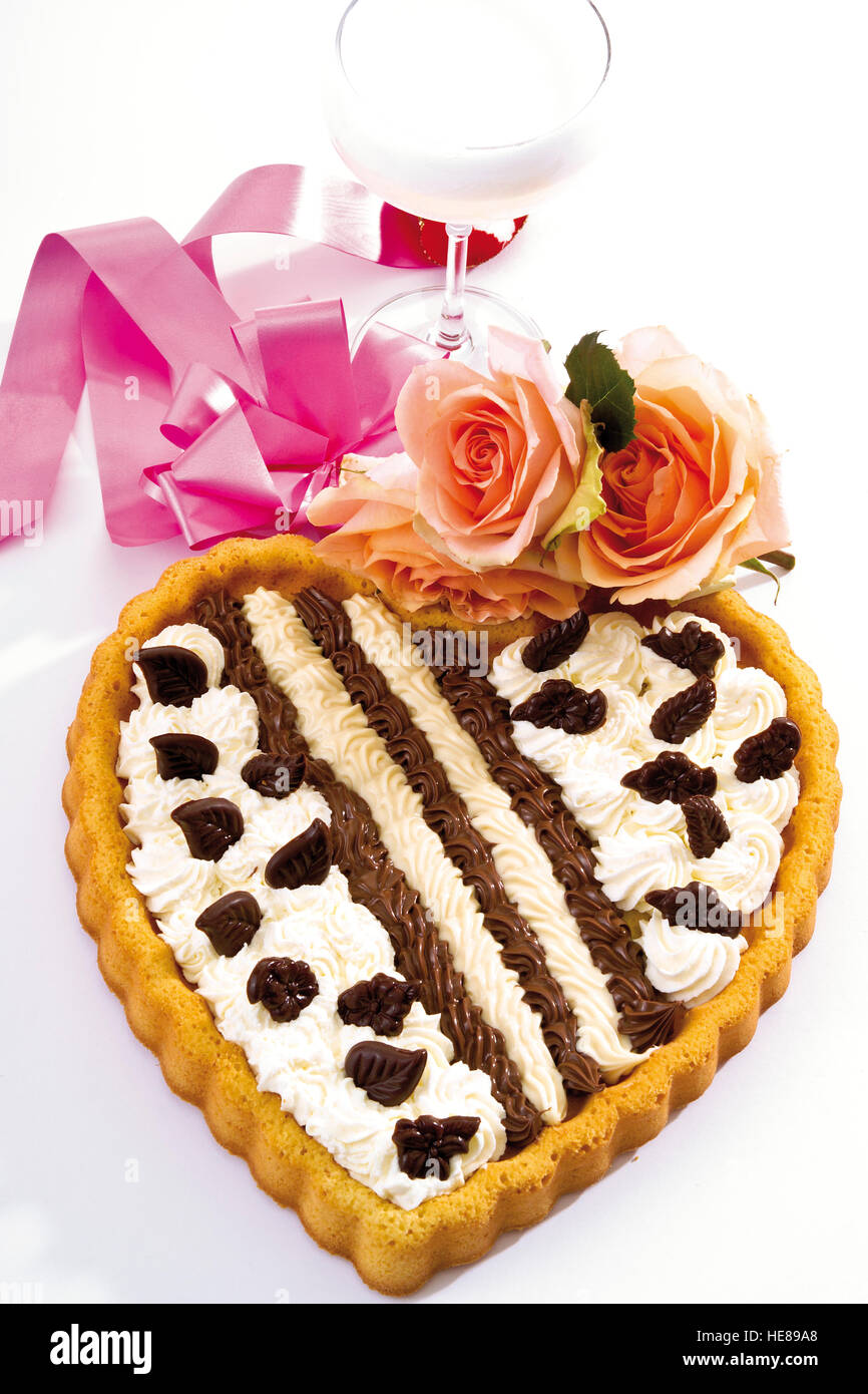 Cream Tart in the shape of a heart adorned with roses in front of a champagne glass and a ribbon Stock Photo