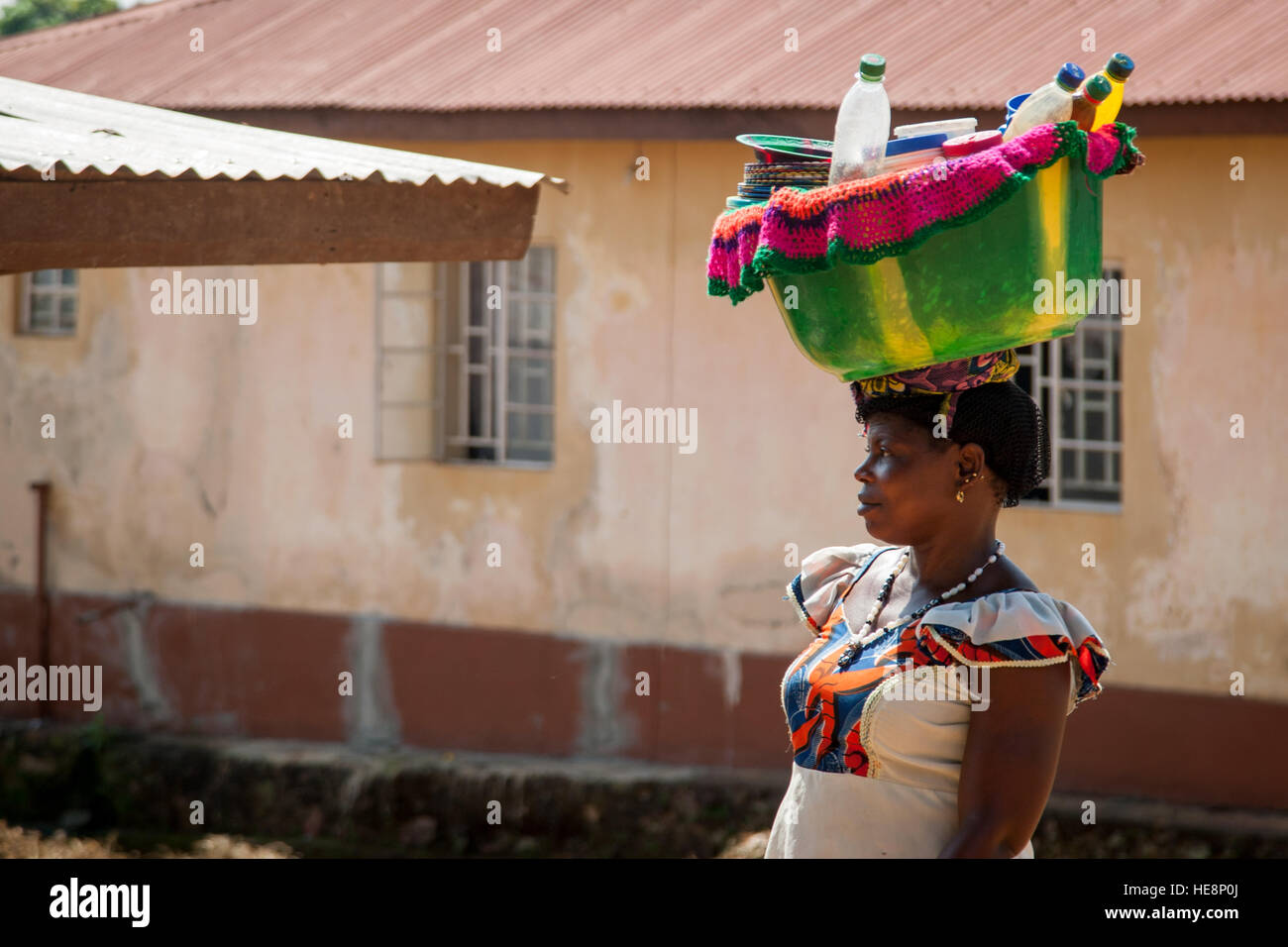 A woman carries a green bowl filled with plastic bottles on her head in front of a plastered house in Kabala, Sierra Leone. Stock Photo