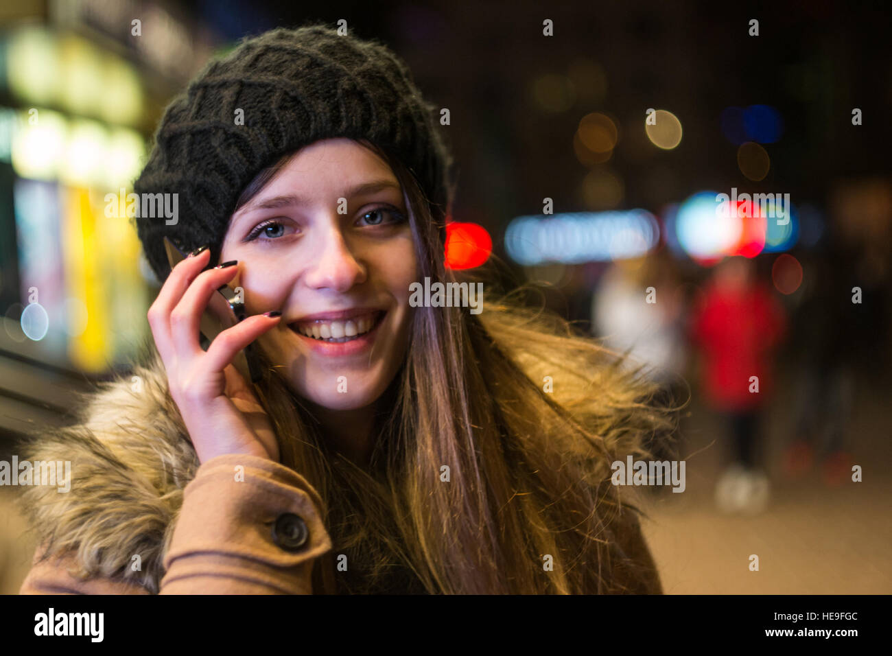 Young happy woman in hat and coat talking on mobile phone smiling looking at camera against night city lights in winter Stock Photo