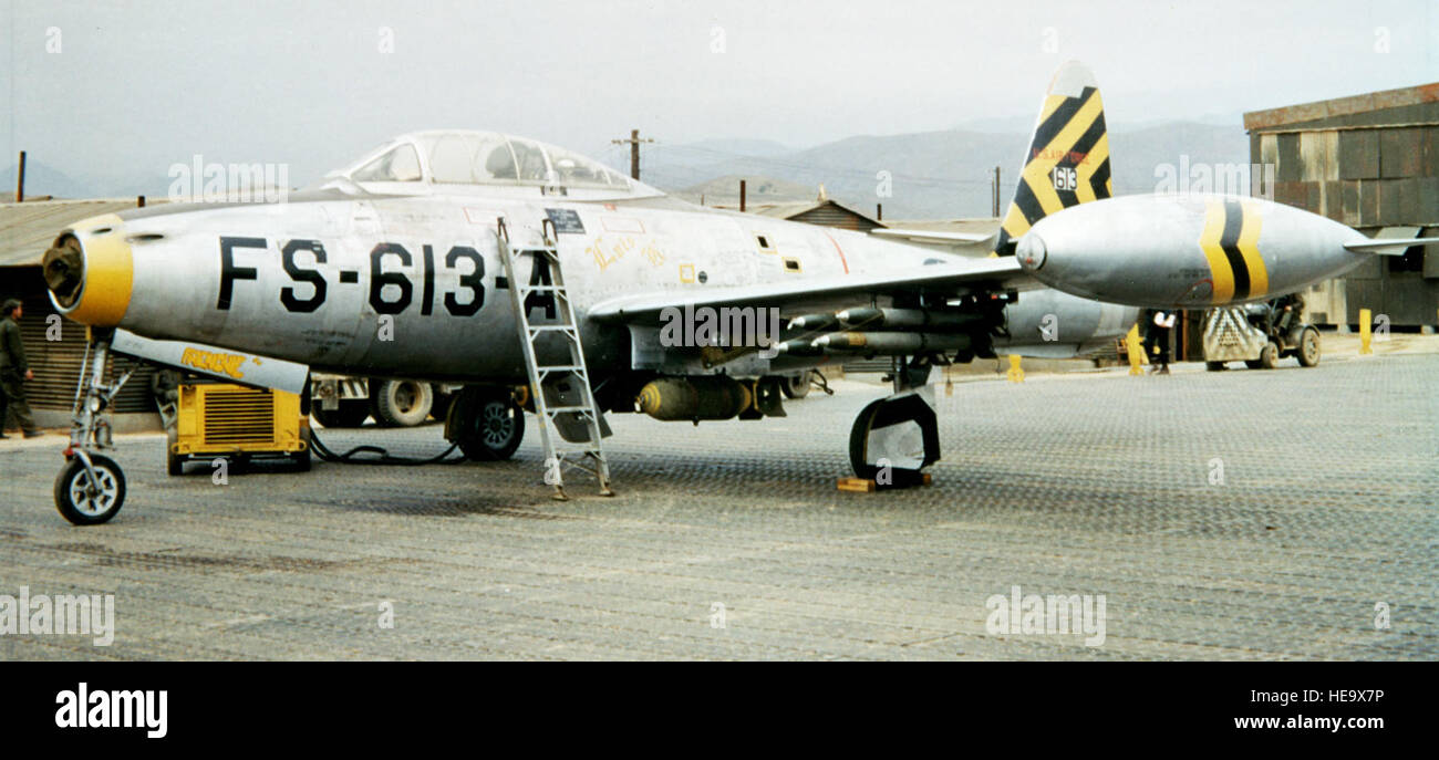 8th FBS/49th FBG.  One-half, left-front view of a Republic F-84E Thunderjet (s/n 51-613) of the 8th Fighter Bomber Squadron (FBS), 49th Fighter Bomber Group (FBG), fitted with external fuel tanks, a bomb, and four rockets, somewhere in Korea.  The three tip tank stripes identify the pilot of this aircraft as squadron leader.  A ladder is propped against the side of the aircraft, leading up to the cockpit.  In the background, a ground crewman is visible, as are some buildings and equipment.  K 90445. Stock Photo