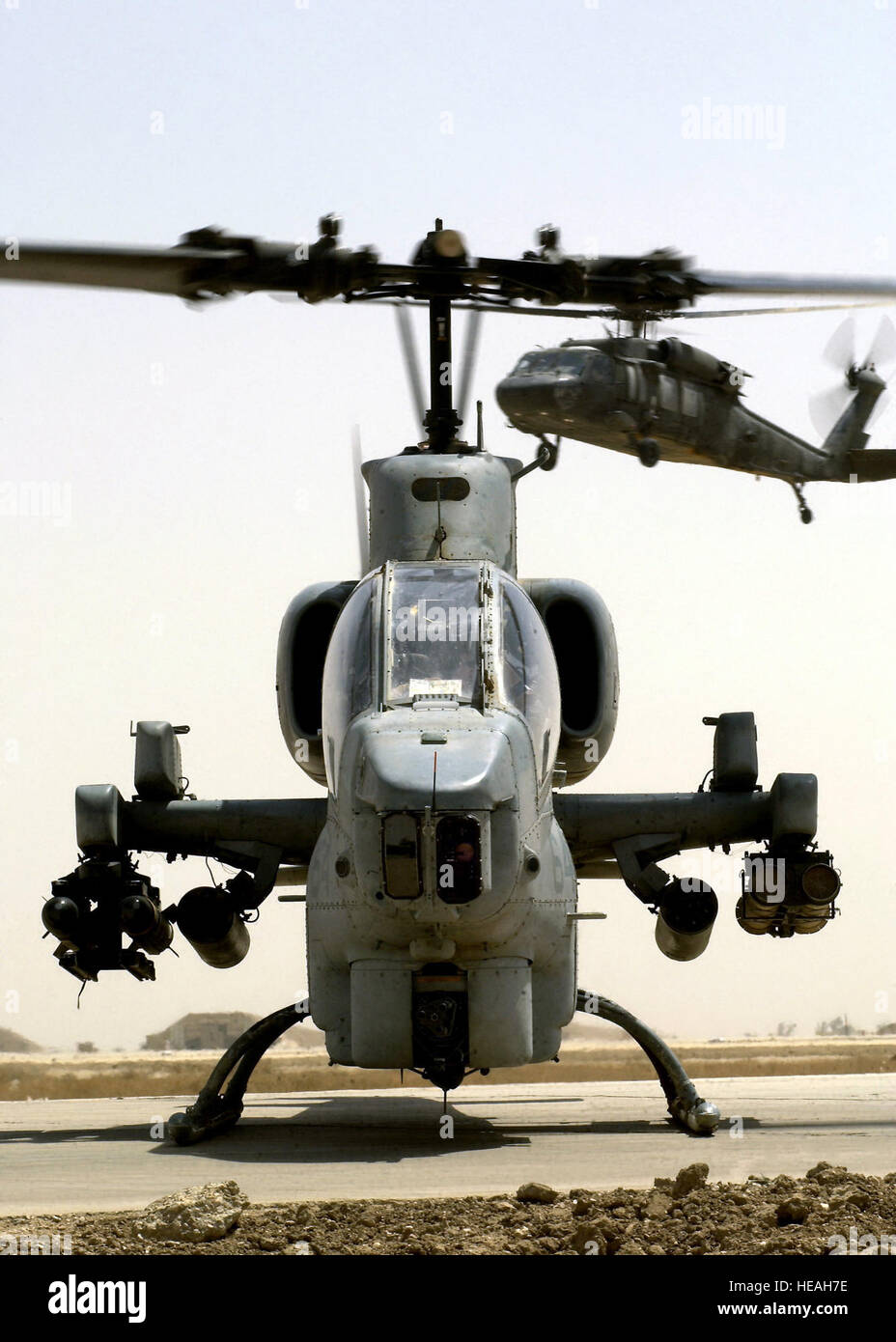 A US Marine Corps (USMC) AH-1W Super Cobra helicopter armed with AGM-114 Hellfire missiles and 2.75-inch Folding Fin Aerial Rockets (FFAR) awaits refueling at a Forward Area Refueling Point (FARP), at Tallil Air Base, Iraq during Operation IRAQI FREEDOM. A US Army (USA) UH-60 Black Hawk helicopter is on approach in the background. Stock Photo