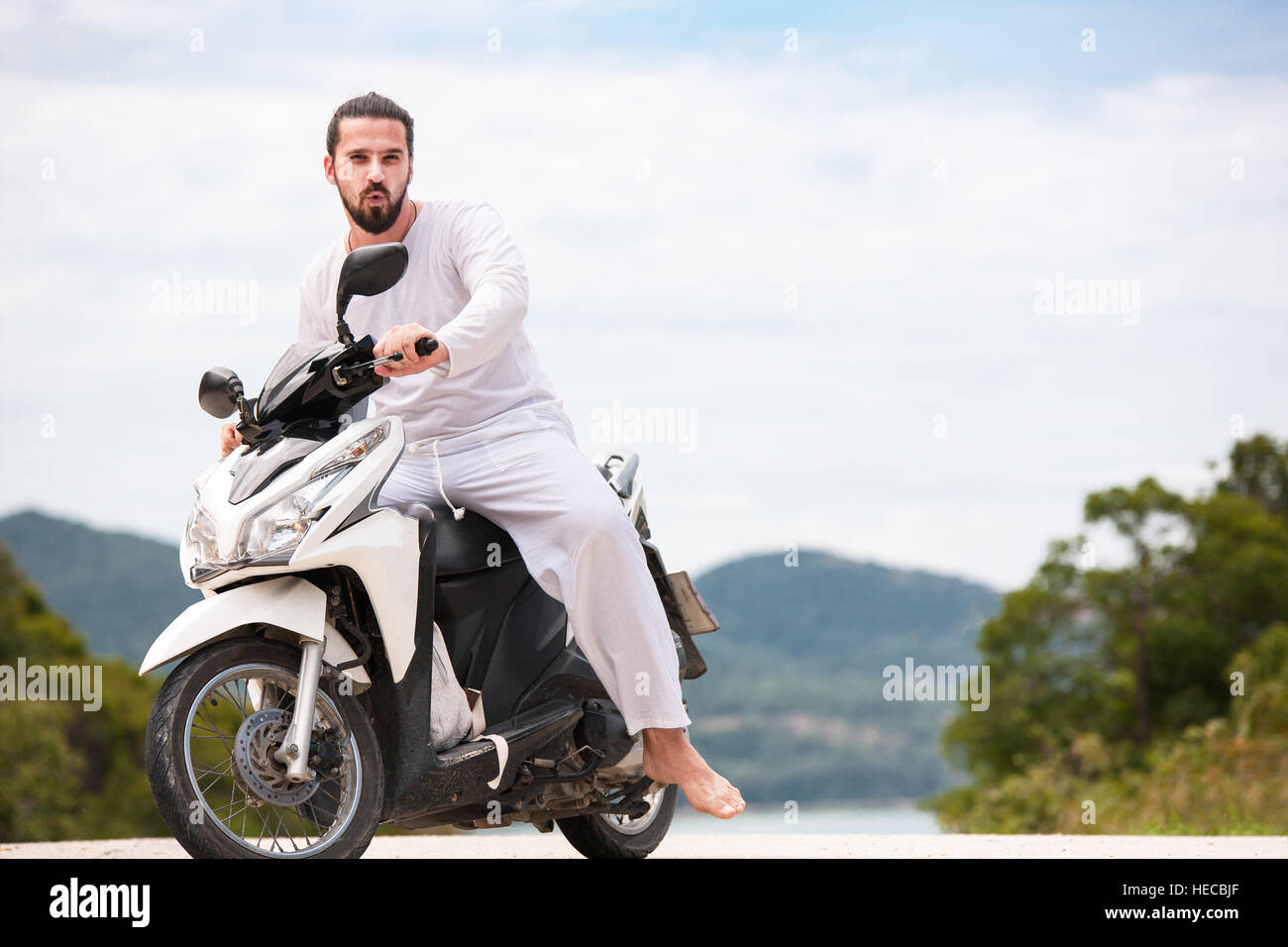 Brutal biker with beard in white sitting on motorbike. Sunny day in the mountains. Stock Photo