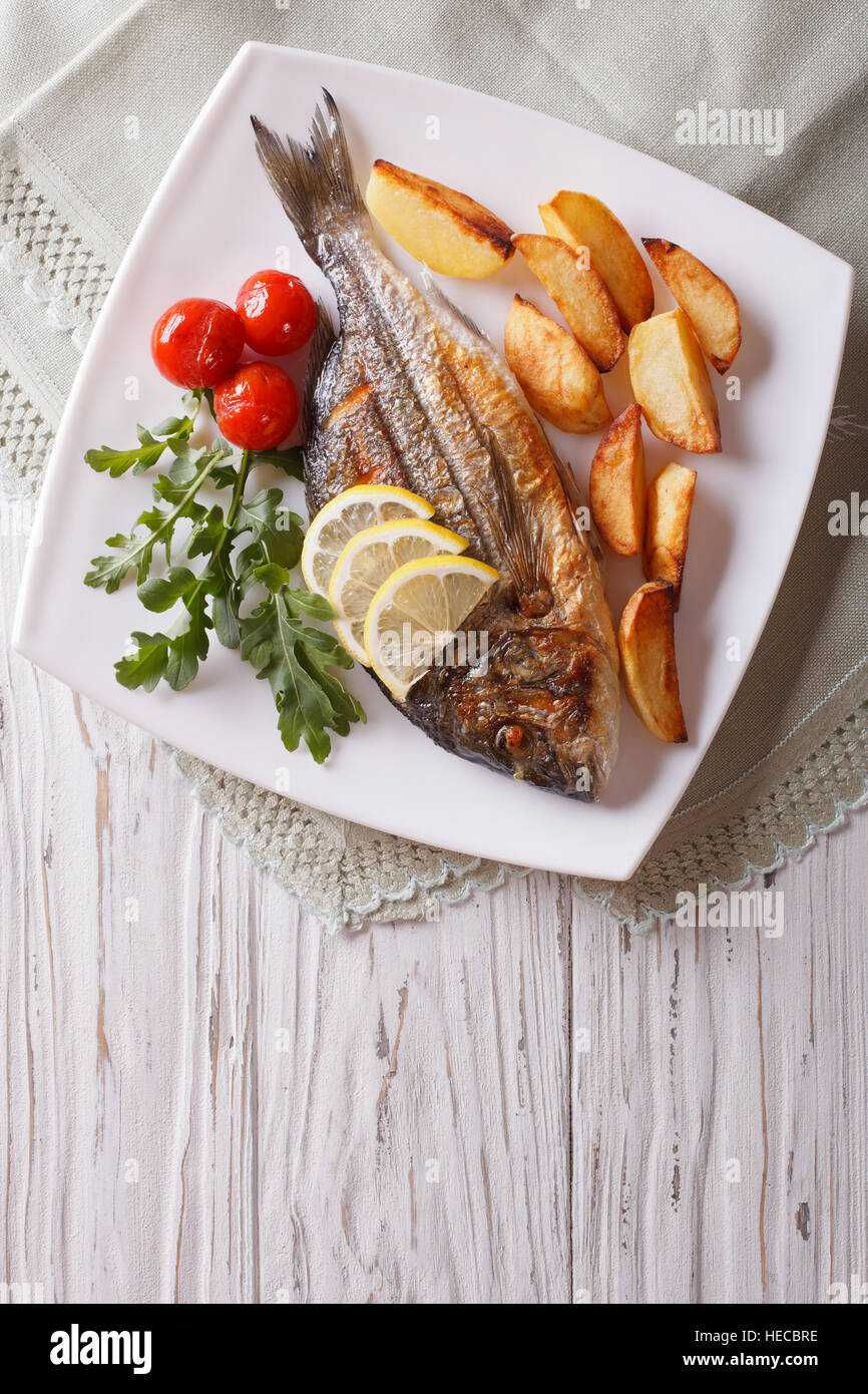 Grilled dorado fish with fried potatoes, lemon and tomato close-up on a plate. Vertical top view Stock Photo