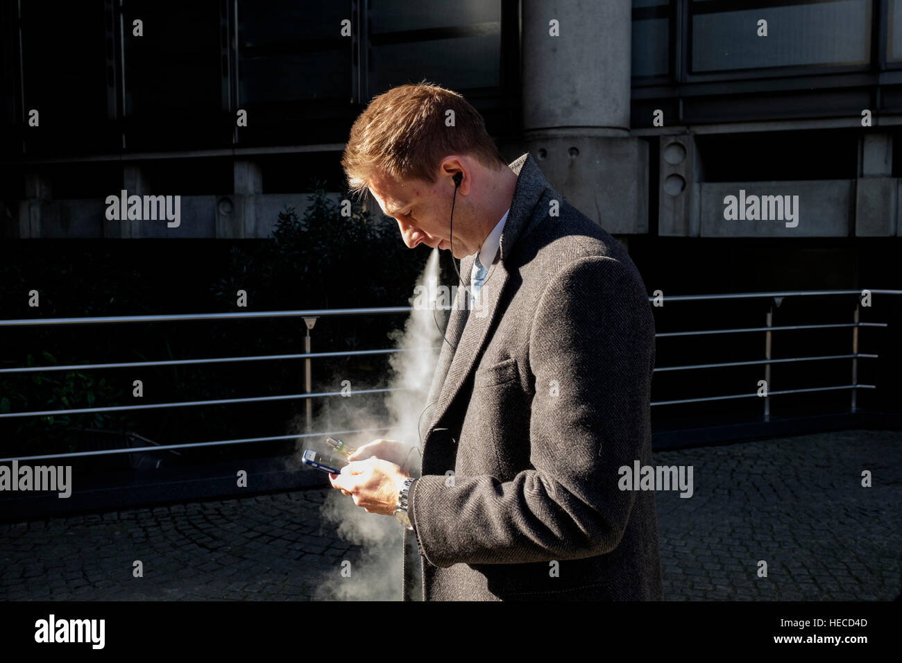 Businessman in the city of London engrossed in his mobile phone enjoys an E-Cigarette Stock Photo