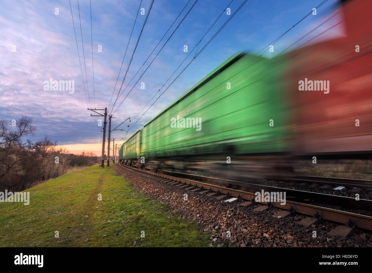 Railway station with cargo wagons and train in motion against blue sky at sunset. Colorful industrial landscape. Railroad Stock Photo