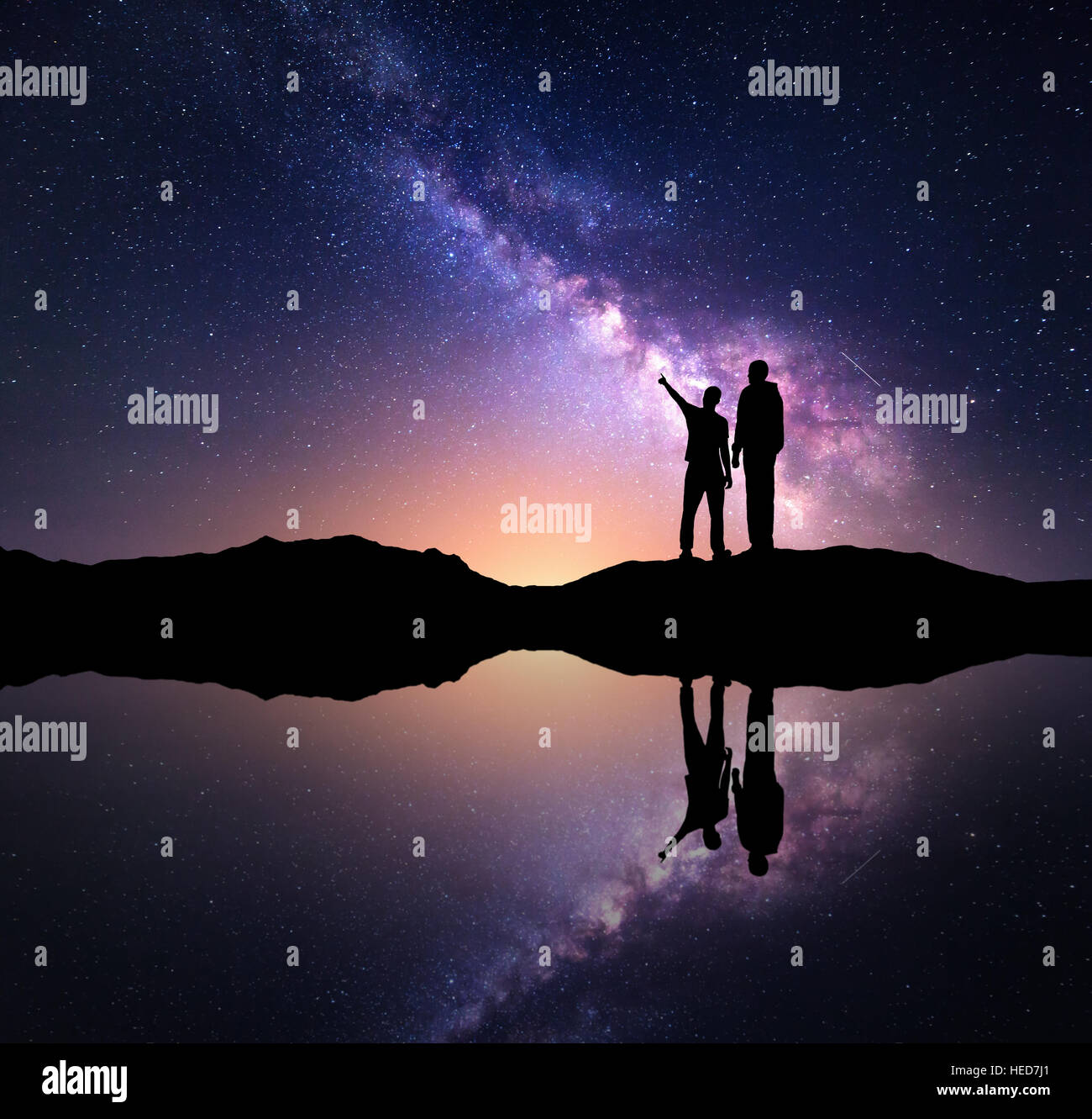Milky Way with silhouette of a family. Father and a son who pointing finger in night starry sky on the mountain near the lake Stock Photo