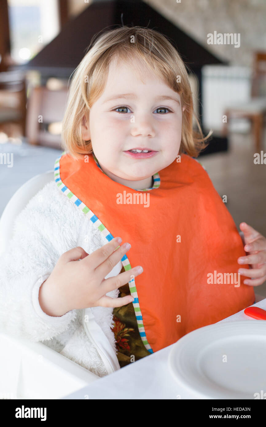 two years old blonde little child with orange bib sitting in table of restaurant looking and
