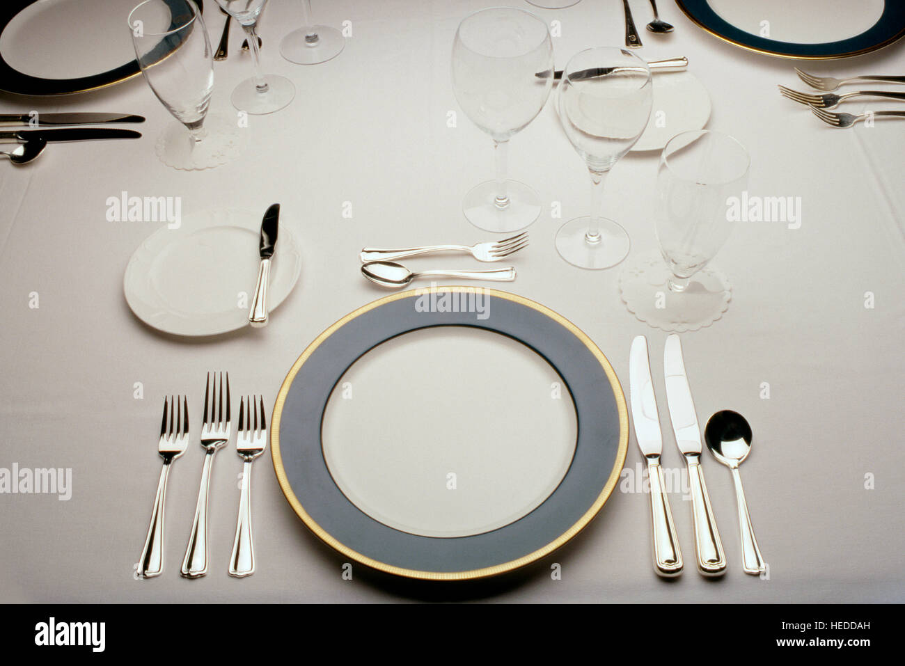 A beautiful, formal dinner setting on a white linen tablecloth. The high end silver service is precisely aligned and polished. Stock Photo