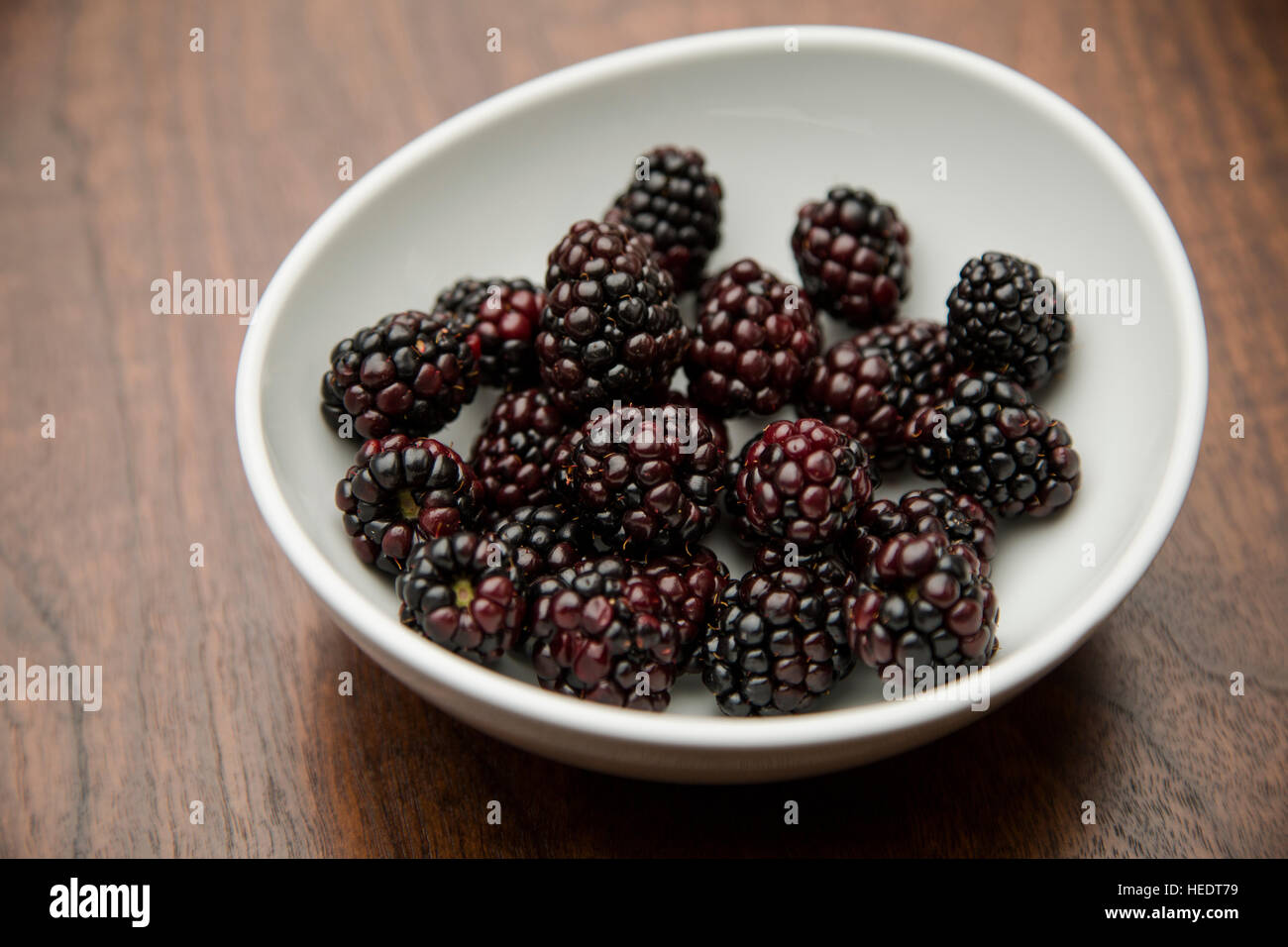 A white ceramic bowl piled with fresh organic blackberries on a dark wood table Stock Photo