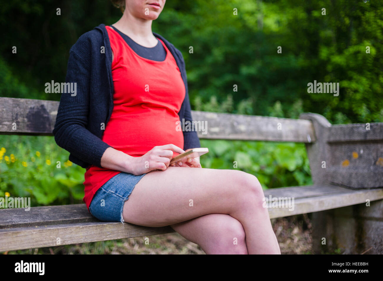 A young pregnant woman is sitting on a bench in the woods on a sunny day and is using her smart phone Stock Photo
