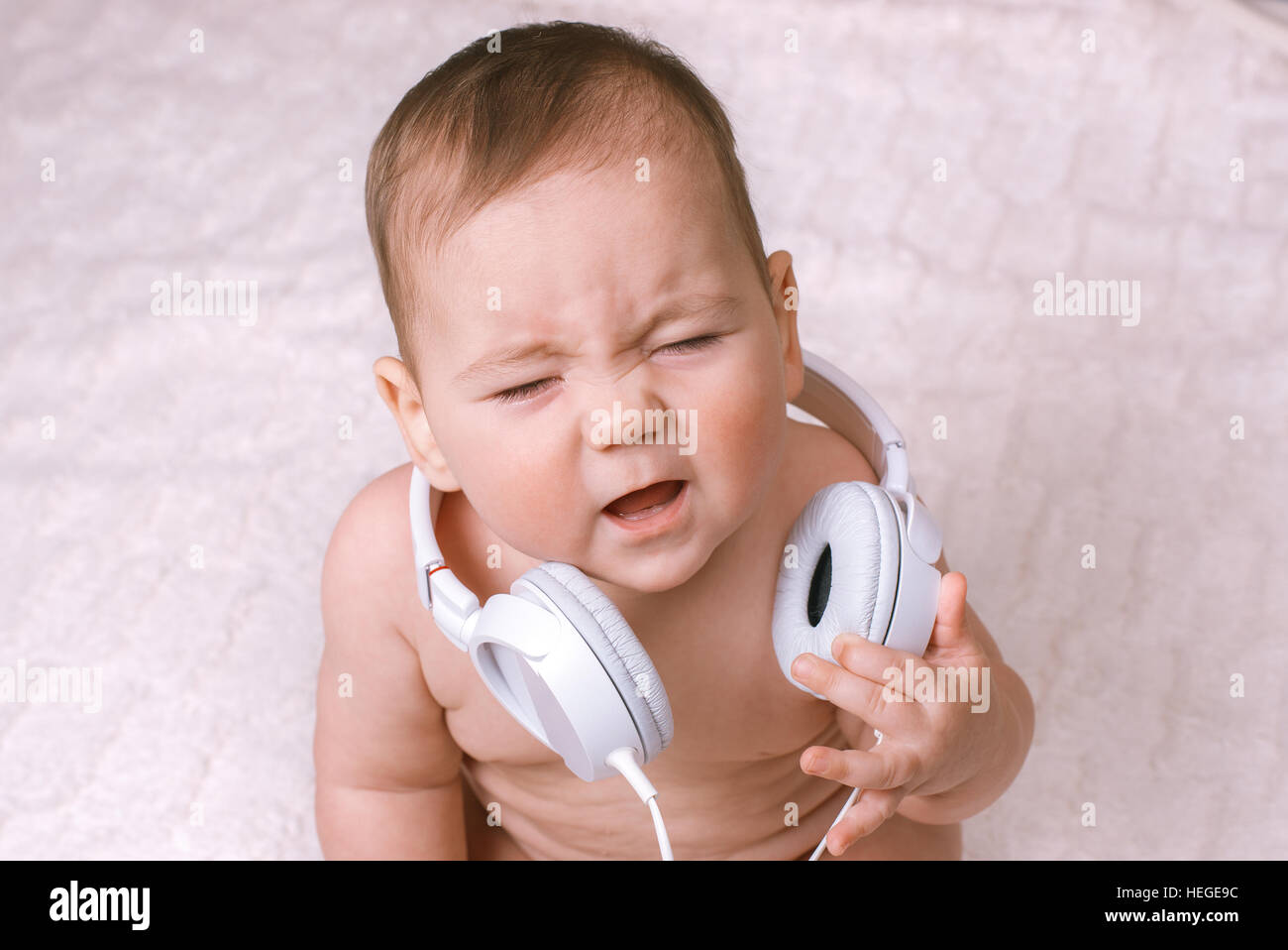 Small baby wearing earphones around its neck as it sits on its woolly rug crying as the audio noise upsets it Stock Photo