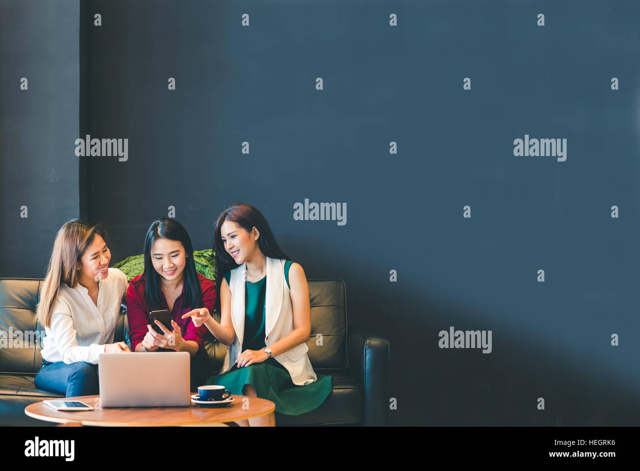 Three beautiful Asian girls using smartphone and laptop, chatting on sofa at cafe with copy space, modern lifestyle with gadget technology or working Stock Photo