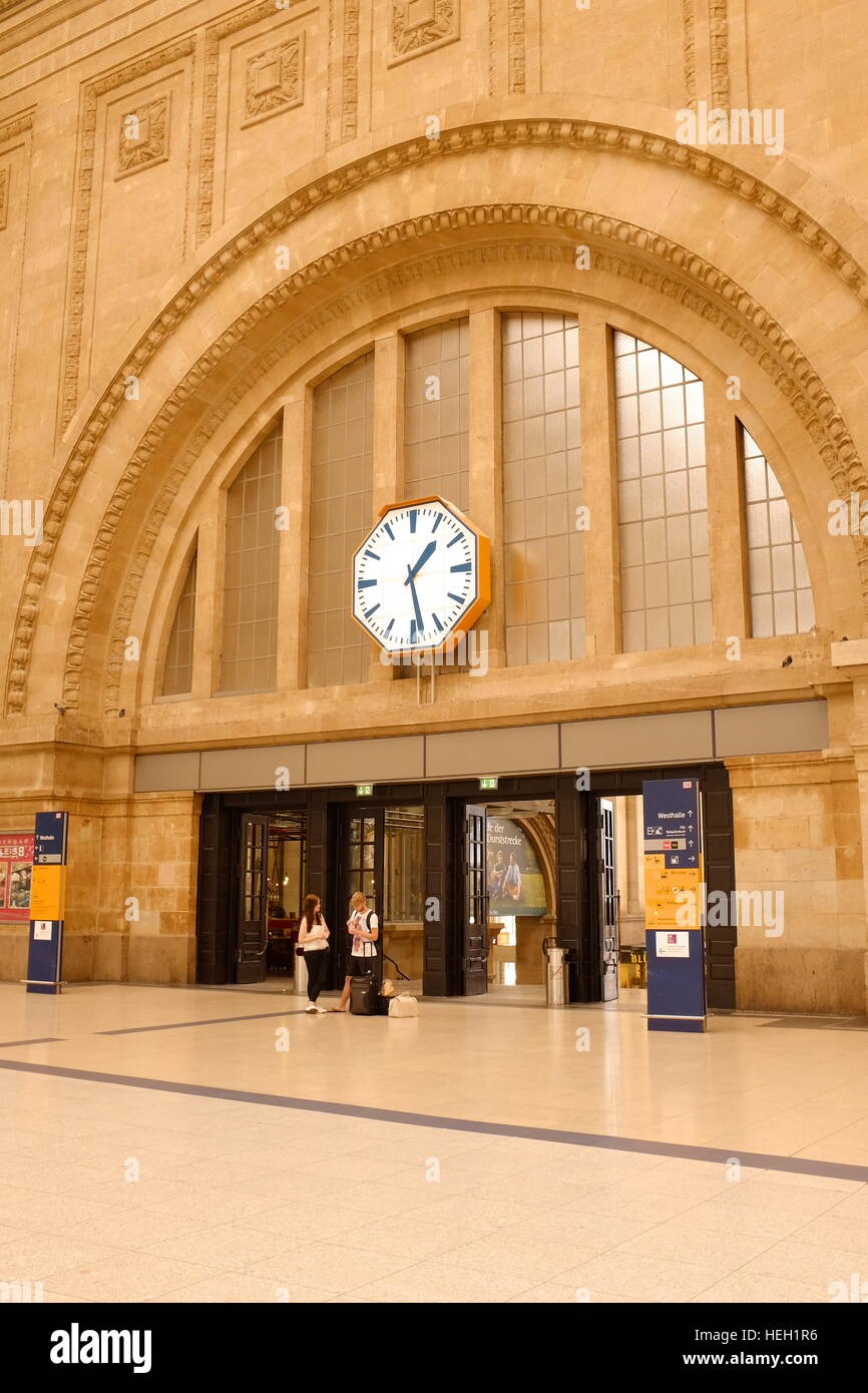 The entrance and foyer in the Leipzig, Germany train station.  A couple converses below a giant clock hanging above them. Stock Photo