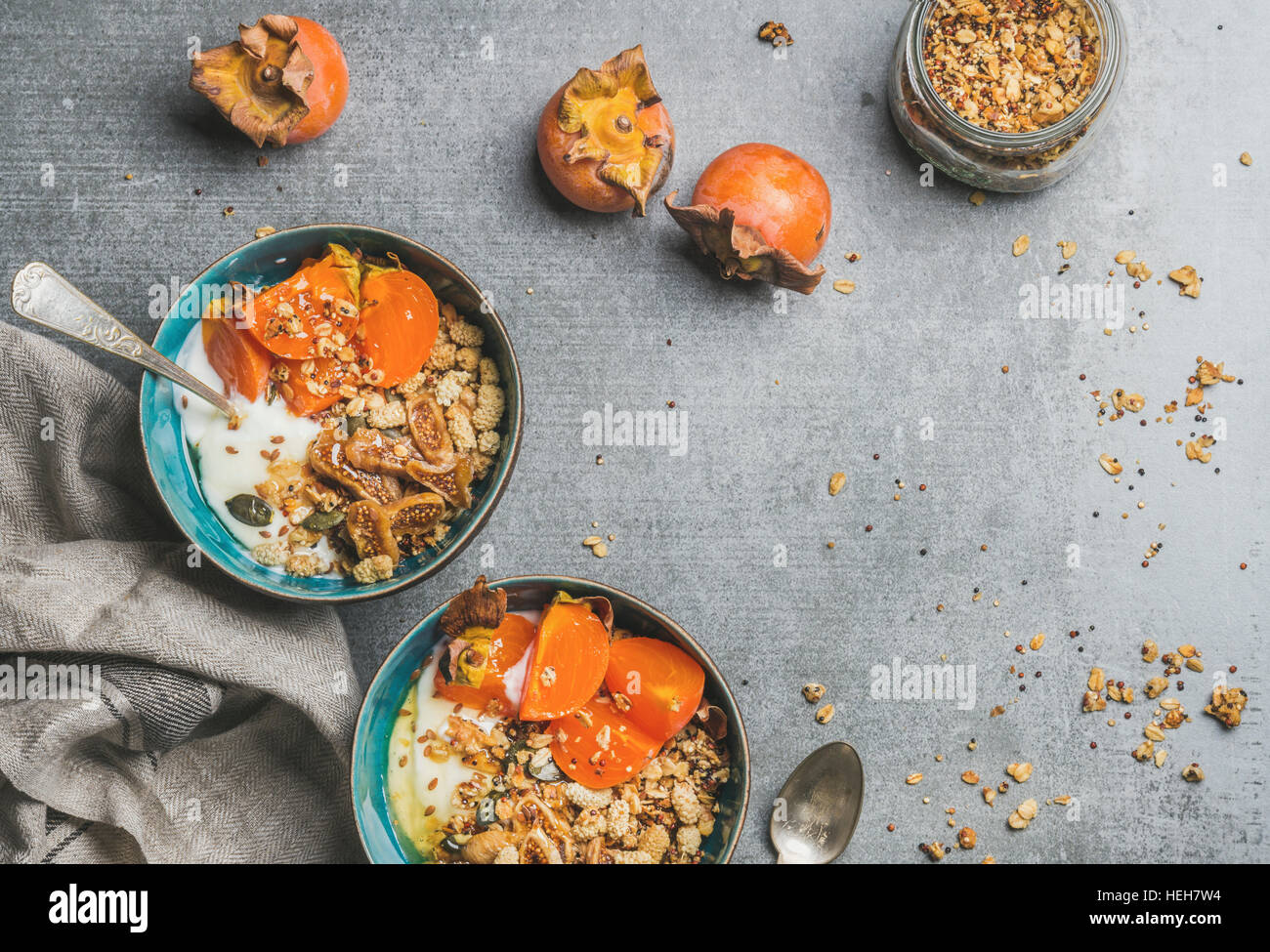 Healthy vegetarian breakfast. Oatmeal, quinoa granola with yogurt, dried fruit, seeds, honey, fresh persimmon in bowls over grey background, top view, Stock Photo