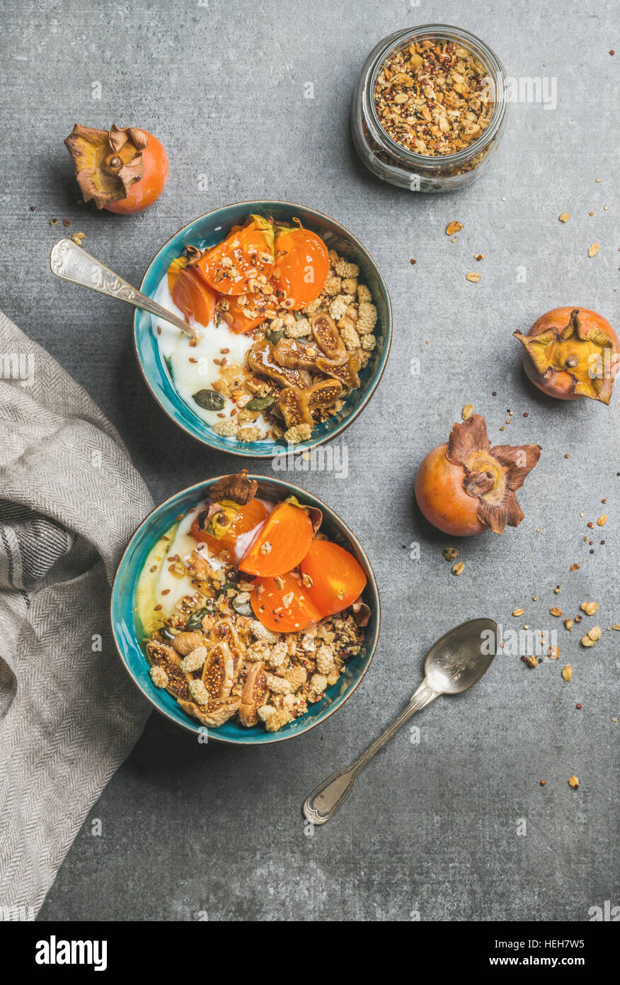 Healthy vegetarian breakfast. Oatmeal, quinoa granola with yogurt, dried fruit, seeds, honey, fresh persimmon in blue ceramic bowls over grey backgrou Stock Photo