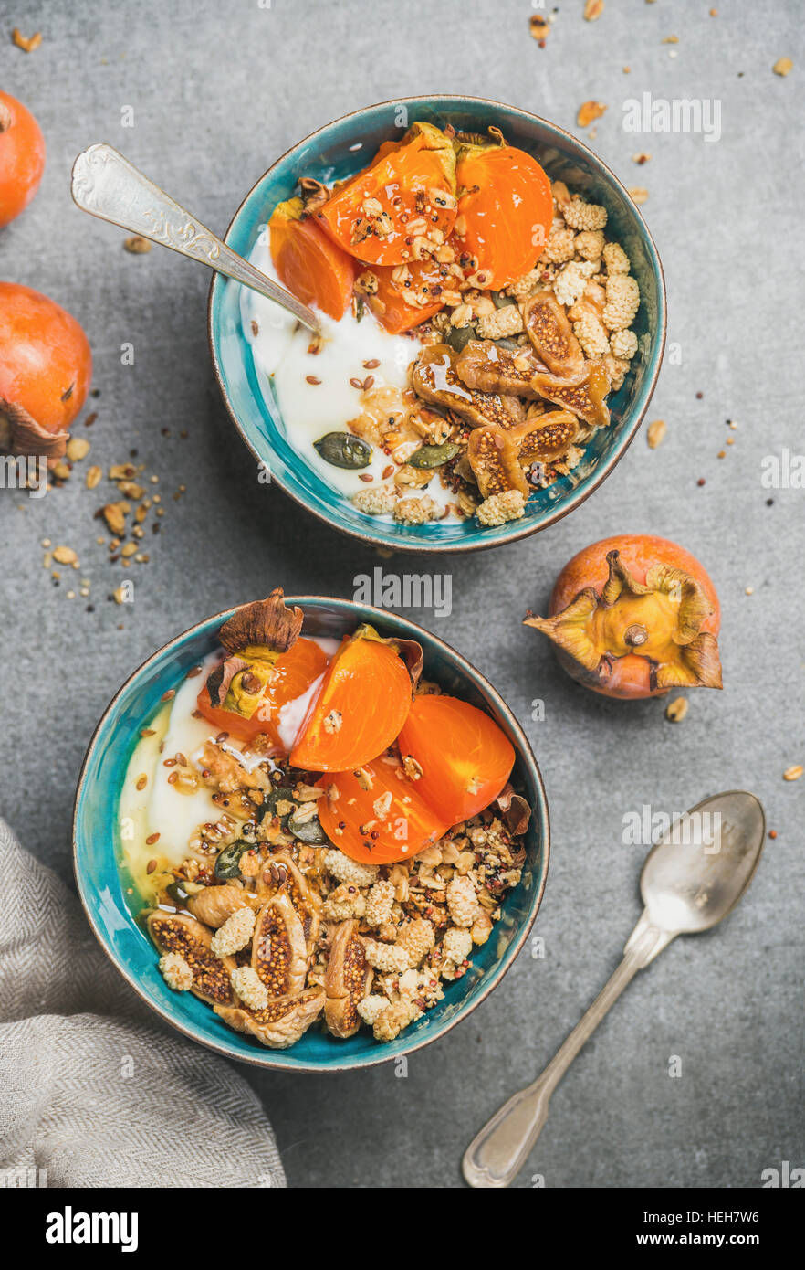 Healthy breakfast. Close-up of oatmeal, quinoa granola with yogurt, dried fruit, seeds, honey, fresh persimmon in blue ceramic bowls over grey backgro Stock Photo
