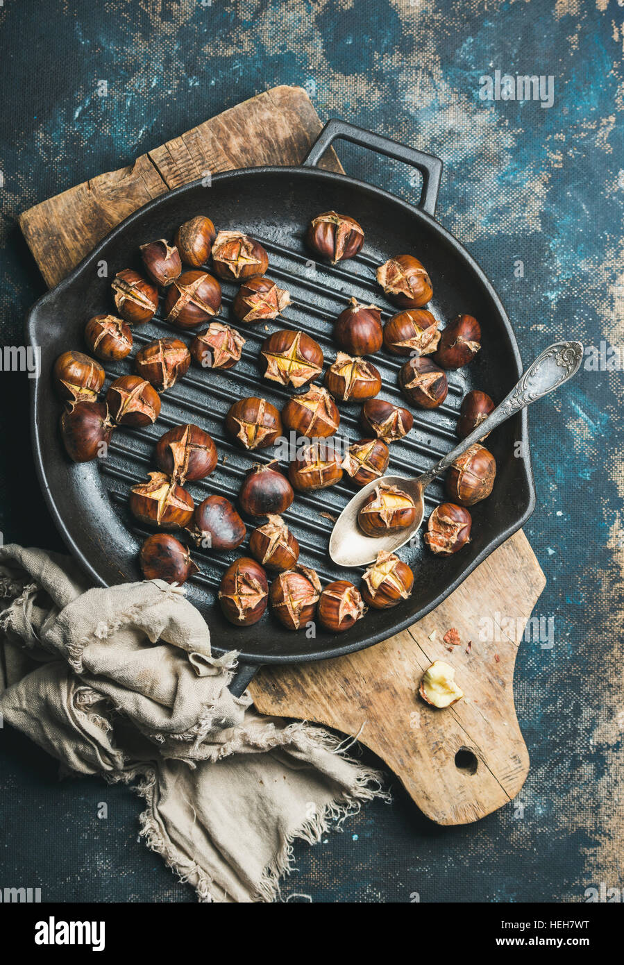 Roasted chestnuts in cast iron grilling pan over rustic wooden board and dark blue shabby plywood background, top view, vertical composition Stock Photo