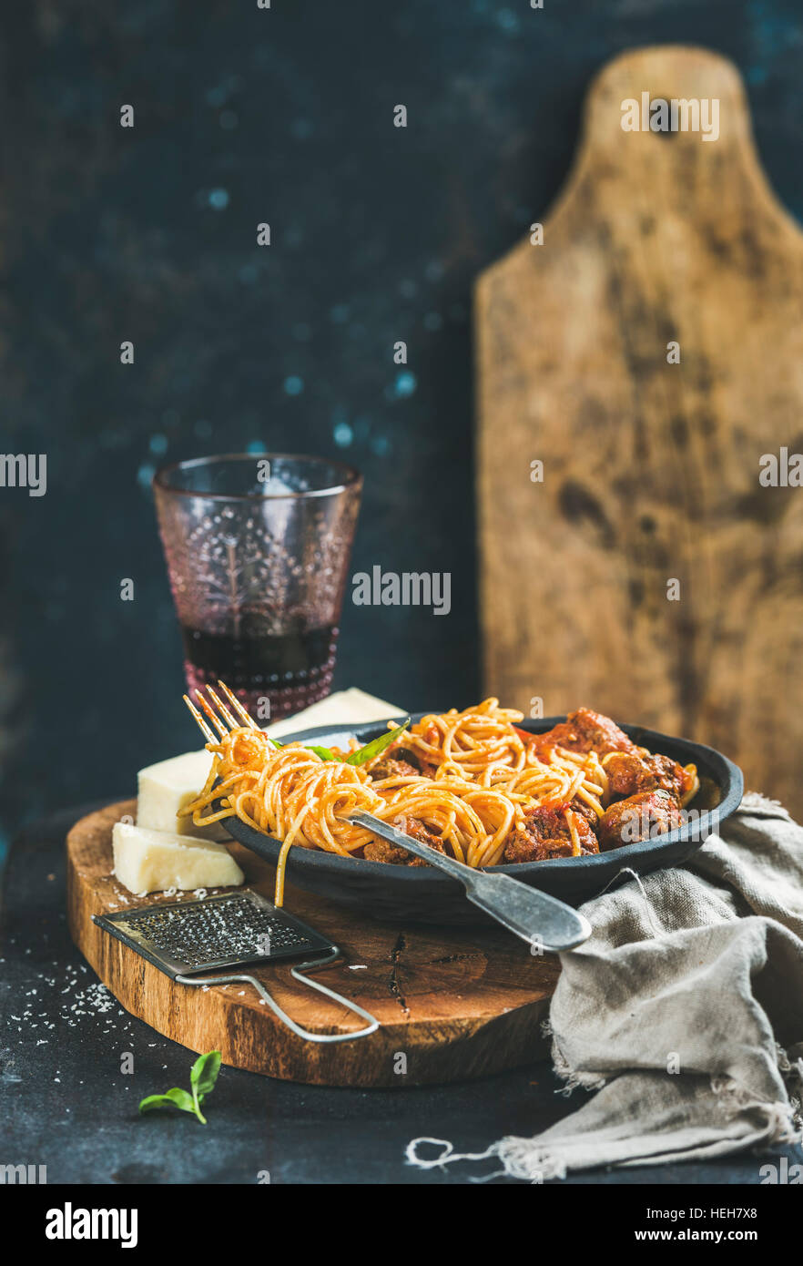 Italian pasta dinner. Spaghetti with meatballas, basil and parmesan cheese in black plate and red wine in glass over rustic wooden board, dark blue wa Stock Photo