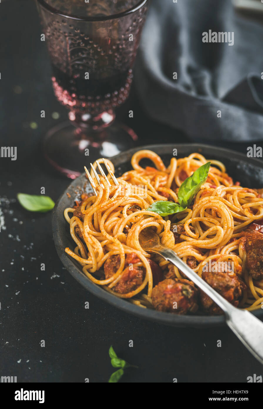 Italian pasta dinner. Spaghetti with meatballas, fresh basil leaves in dark plate and red wine in vintage glass over black background, selective focus Stock Photo