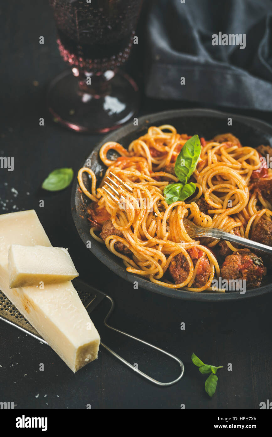 Italian pasta dinner. Spaghetti with meatballas, basil and parmesan cheese in dark plate and red wine in glass over black background, selective focus. Stock Photo