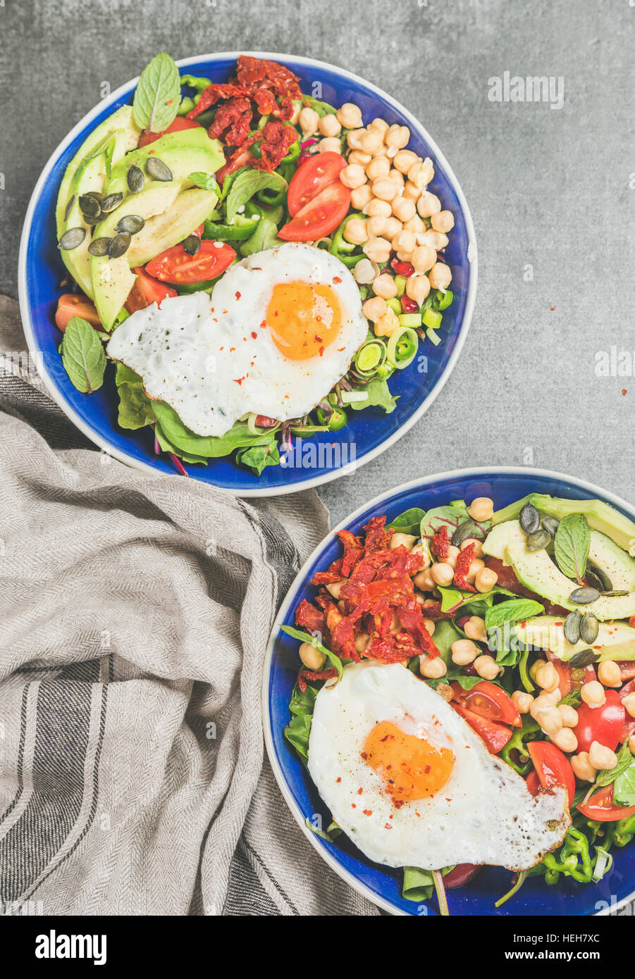 Healthy breakfast bowls with fried egg, chickpea sprouts, seeds, fresh vegetables and greens over grey concrete background, top view. Clean eating, di Stock Photo