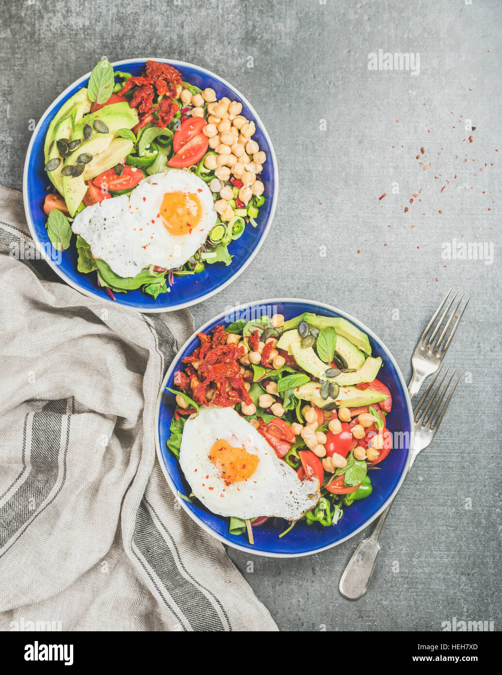 Healthy breakfast bowls with fried egg, chickpea sprouts, seeds, vegetables and greens over grey concrete background, top view, copy space. Clean eati Stock Photo