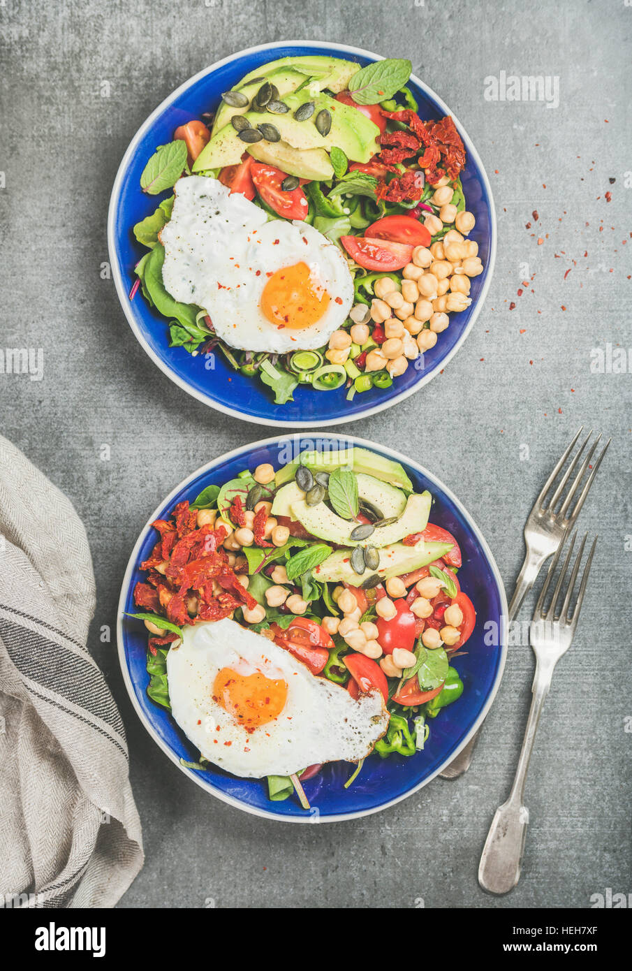 Healthy breakfast with fried egg, chickpea sprouts, seeds, fresh vegetables and greens in blue bowls over grey concrete background, top view. Clean ea Stock Photo