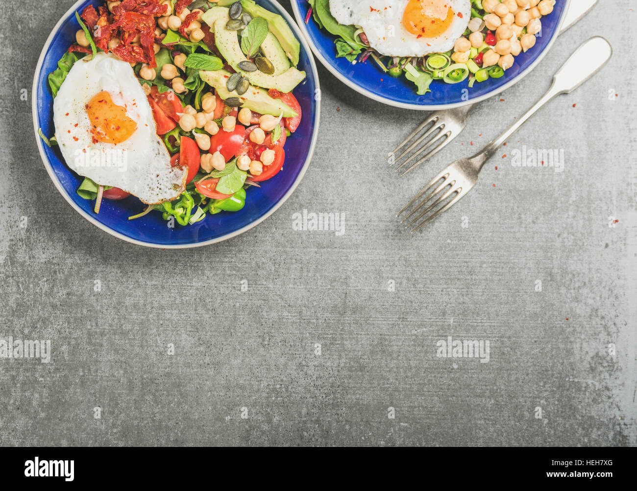 Healthy breakfast with fried egg, chickpea sprouts, seeds, vegetables and greens in blue ceramic bowls over grey concrete background, top view, copy s Stock Photo