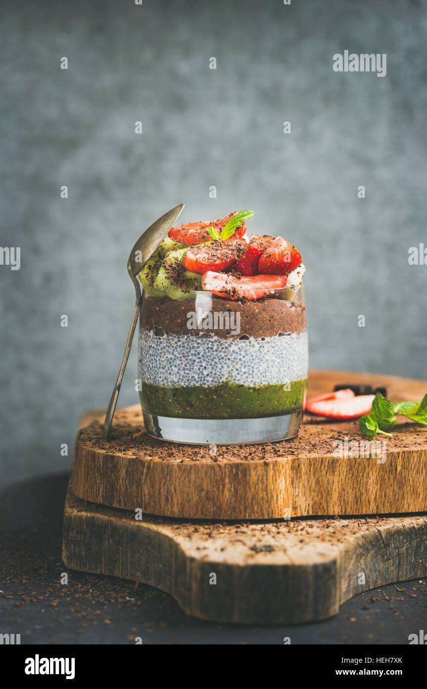 Healthy detox breakfast concept. Matcha, almond milk, cocoa chia seed pudding with fresh fruit, berries, mint, chocolate in glass over wooden board, g Stock Photo