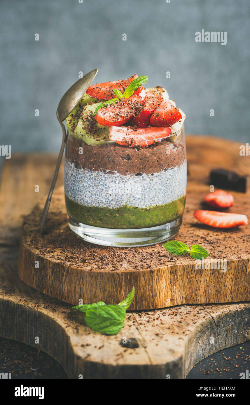 Healthy vegan breakfast concept. Matcha, almond milk, cocoa chia seed pudding with fresh fruit, berries, mint, chocolate in glass over wooden board, g Stock Photo