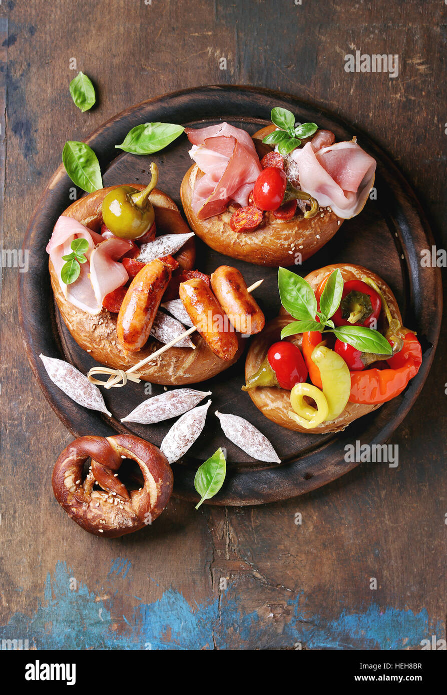 Variety of meat snacks fried sausages, wienerwurst, ham, marinated chili peppers served in salted pretzels with fresh basil on wood chopping board ove Stock Photo