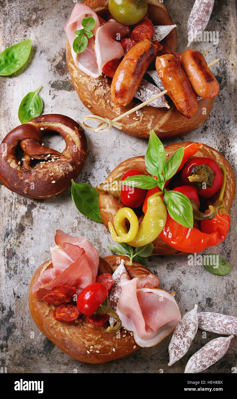 Variety of meat snacks fried sausages, wienerwurst, ham, marinated chili peppers served in salted pretzels with fresh basil over old metal background. Stock Photo