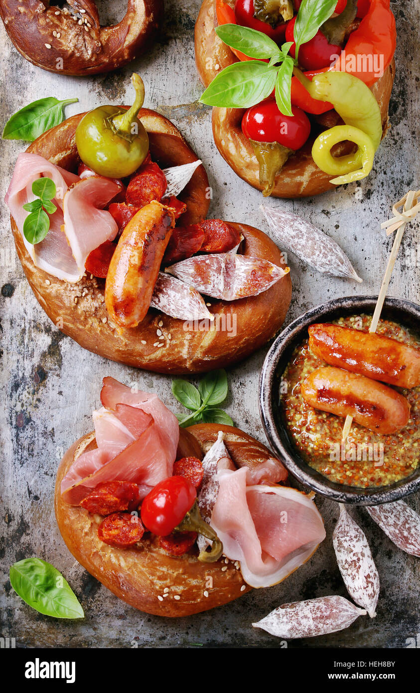 Variety of meat snacks fried sausages, wienerwurst, ham, marinated chili peppers served in salted pretzels with fresh basil and mustard over old metal Stock Photo