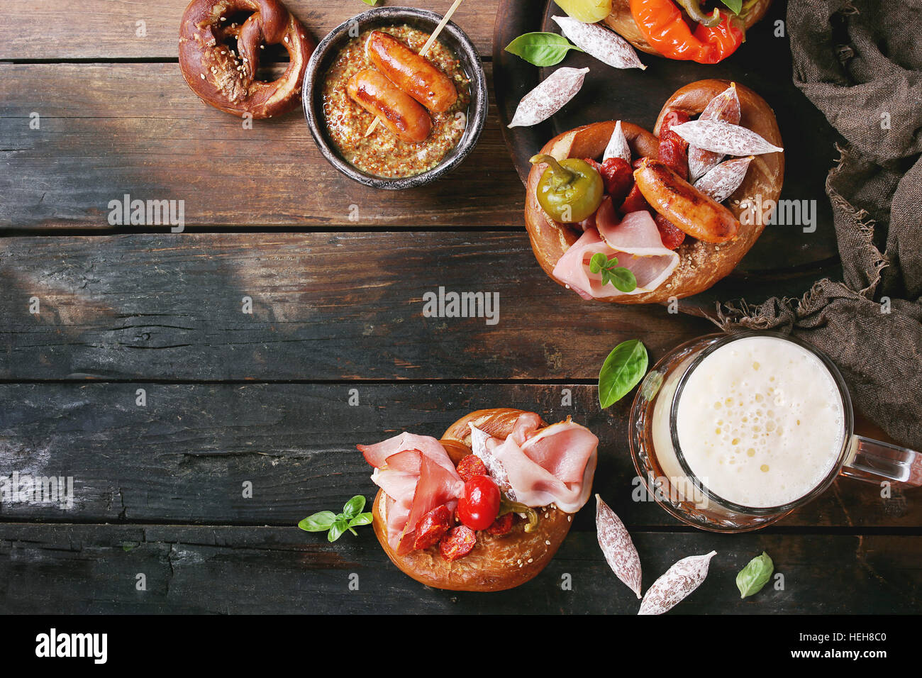 Variety of meat snacks fried sausages, wienerwurst, ham, marinated chili peppers served in salted pretzels with fresh basil and glass of lager beer ov Stock Photo