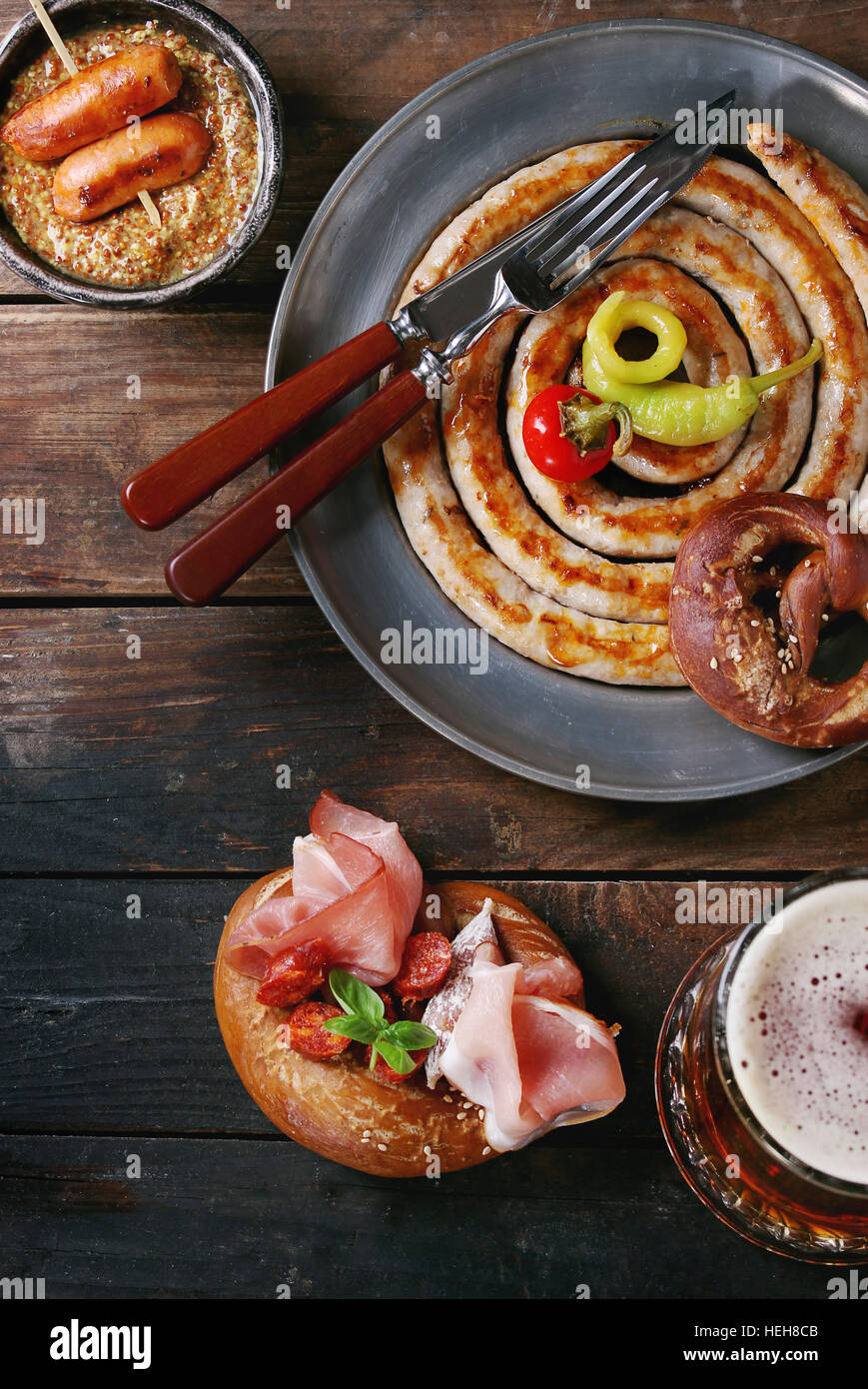 Big spiral fried sausage, meat snacks wienerwurst, ham, marinated chili peppers served in salted pretzels and plate with glass of lager beer and musta Stock Photo