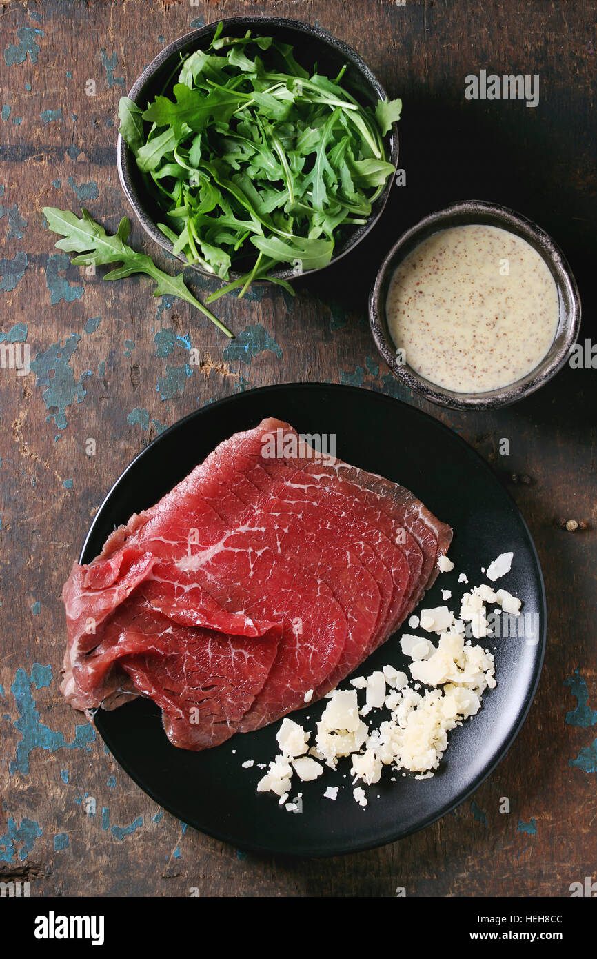 Ingredients for making carpaccio. Raw sliced beef on black plate, mustard and parmesan sauce, cheese, bowl of arugula over old dark texture background Stock Photo