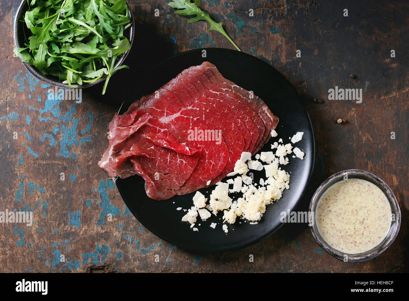 Ingredients for making carpaccio. Raw sliced beef on black plate, mustard and parmesan sauce, cheese, bowl of arugula over old dark texture background Stock Photo
