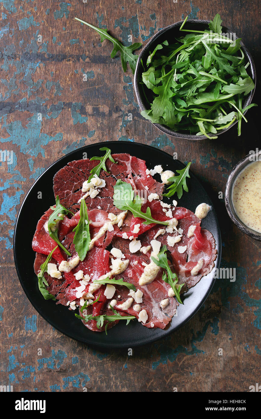 Beef carpaccio on black plate with mustard and parmesan sauce, cheese and arugula over old dark texture background. Top view with space for text Stock Photo