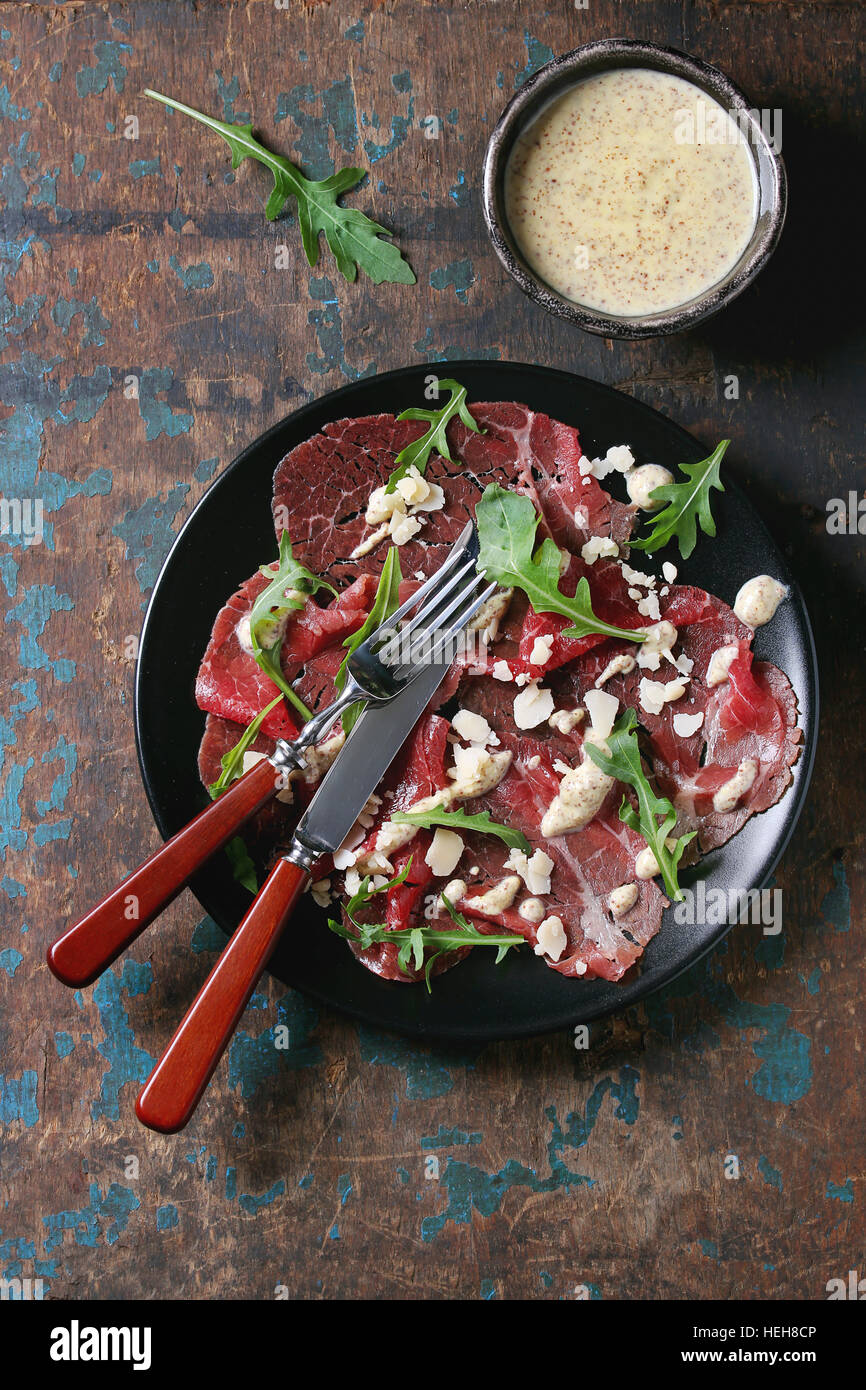 Beef carpaccio on black plate with mustard and parmesan sauce, cheese and arugula, served with fork, knife and ingredients above over old dark texture Stock Photo