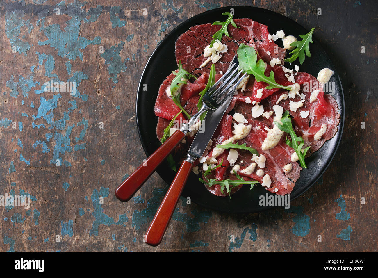 Beef carpaccio on black plate with mustard and parmesan sauce, cheese and arugula, served with fork and knife over old dark texture background. Top vi Stock Photo