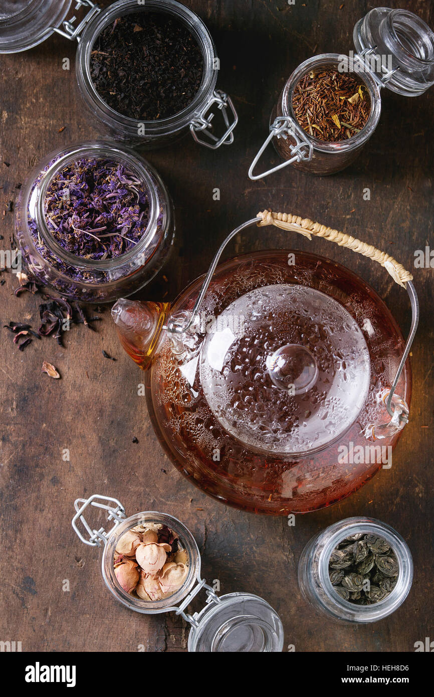 Variety of black, green and herbal dry tea leaves in glass jars with vintage strainer and teapot of hot tea over old dark wooden background. Top view Stock Photo