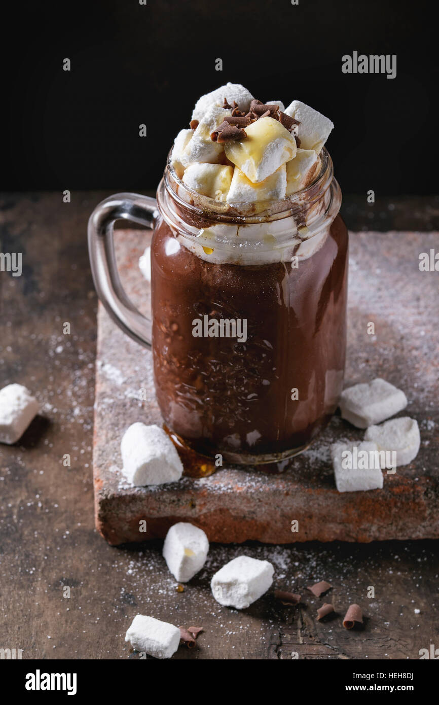 Hot chocolate with homemade marshmallow, chocolate chips and syrup in mason jar, standing on terracotta board over old dark wooden background. Space f Stock Photo