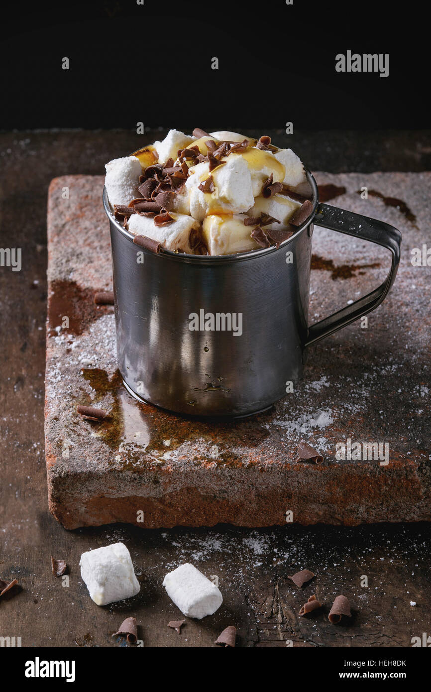 Hot chocolate with homemade marshmallow, chocolate chips and syrup in metal mug, standing on terracotta board over old dark wooden background. Copy sp Stock Photo
