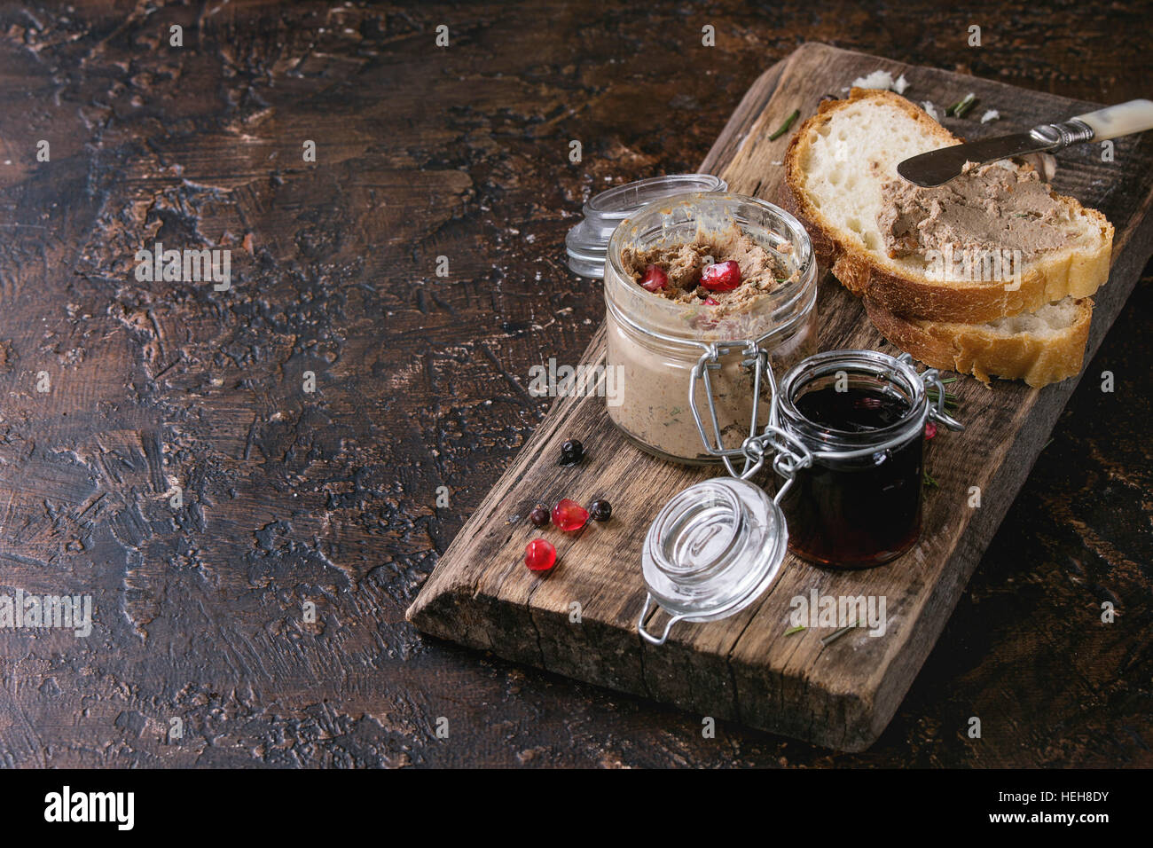 Glass jars of chicken liver pate with blackcurrant jam, pomegranate grain and sliced bread, served with vintage knife on wooden chopping board over da Stock Photo