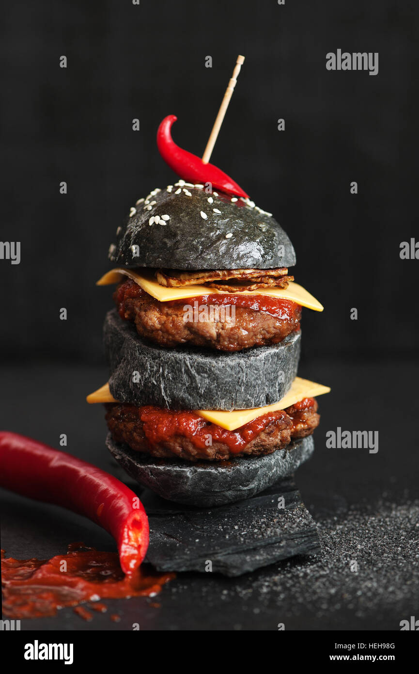 Black burger with meat, cheese, fried onion, tomato sauce and hot chili pepper on the black table Stock Photo