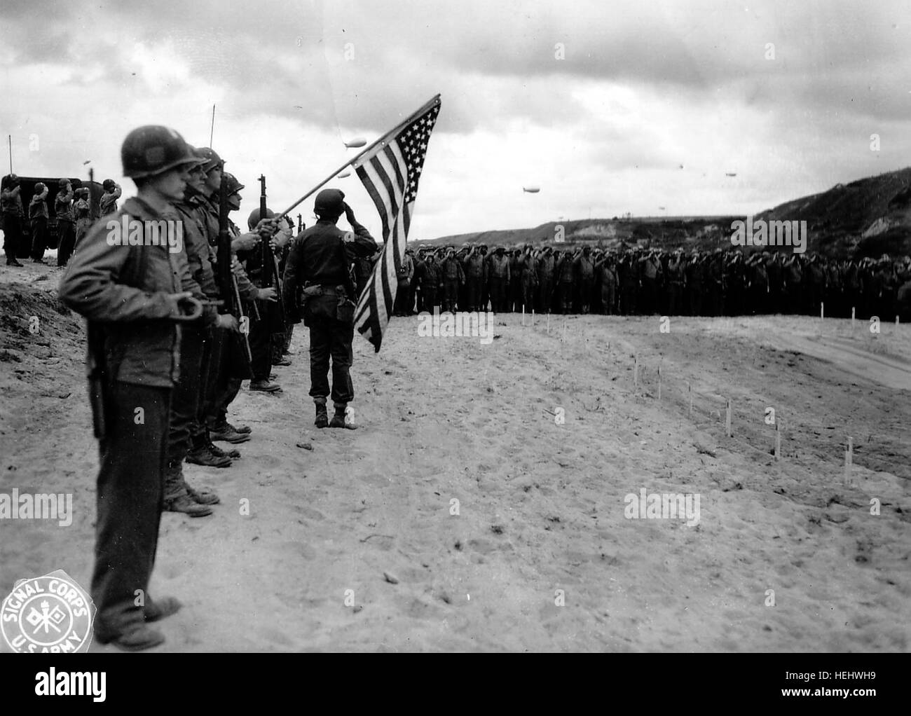 La garde au drapeau et le clairon, une messe à l'emplacement du premier cimetière provisoire à Vierville, Voir ici : http://omahabeach.vierville.free.fr/8431-CimetiereProvisoireVierville.html On distingue les piquets en bois au-dessus de chaque tombe. Plusieurs ballons de barrage dans le ciel. Deux films de cette cérémonie: http://www.criticalpast.com/video/65675060423 Military-Chaplain funeral-services graves-of-dead-United-States-soldiers Eucharist http://www.criticalpast.com/video/65675061293 United-States-soldiers American-cemetery burial-ceremony 3-volley-rifle-salute 4 photos de cette me Stock Photo