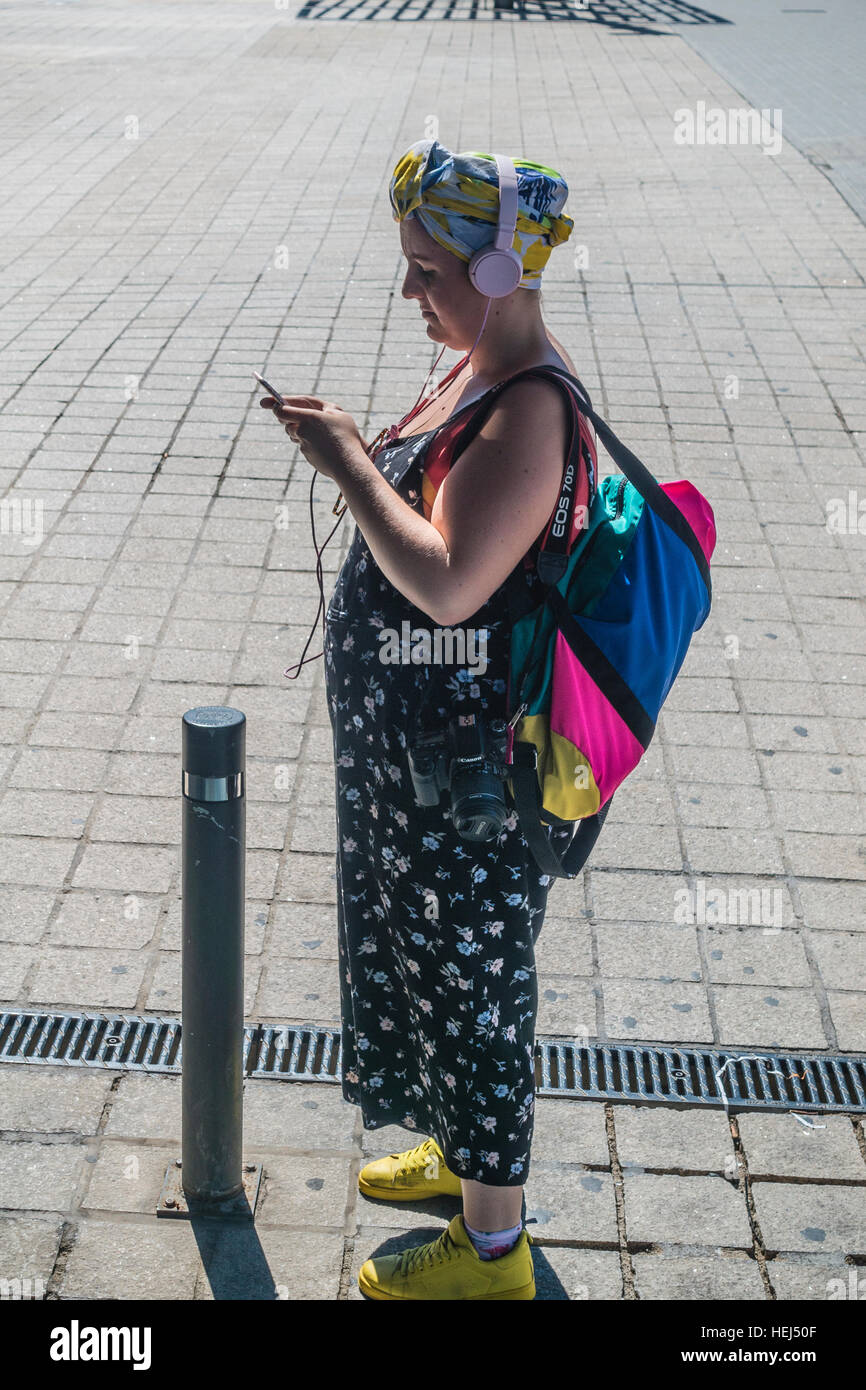 Female, high-tech tourist with a camera, backpack, headphones, and smartphone, reads as she stands on a sidewalk in Barcelona, Stock Photo