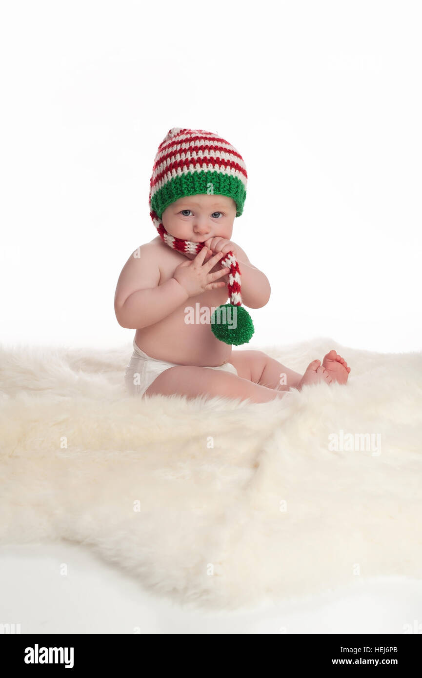 A seven month old baby boy sitting on a sheepskin rug. He is wearing a long, red, white and green striped stocking cap and chewing on the hat's tail.  Stock Photo
